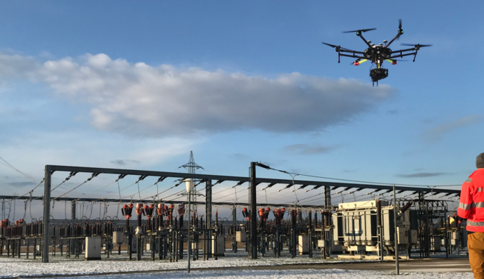 Industrie-Drohne fliegt über Umspannwerk zur Inspektion, gesteuert von einem Techniker in Warnkleidung.